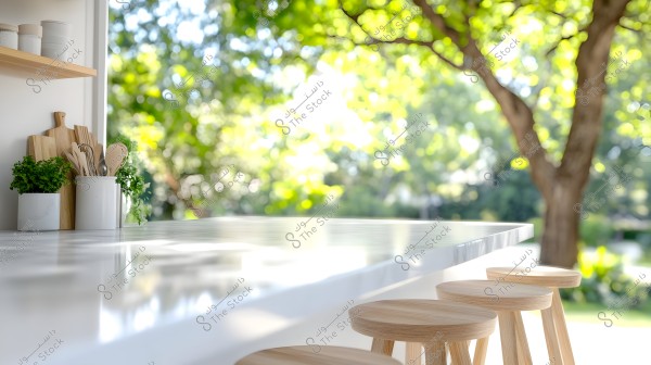 An image of an open kitchen facing an outdoor garden. The picture features a shiny marble countertop surrounded by wooden stools on the right. To the left, there are potted plants and wooden kitchen utensils placed in a white container, with wooden cutting boards behind them. In the background, green trees are brightly lit by sunlight.