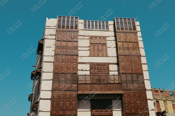 A traditional building showcasing Hijazi architectural style, featuring a facade adorned with intricate wooden work of balconies and mashrabiyas. The warm wood tones contrast with the clear blue sky in the background.
