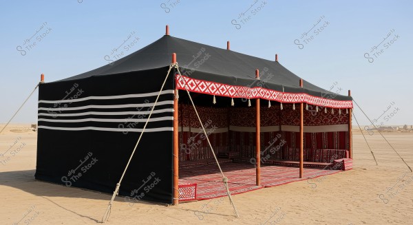 An image of a traditional Bedouin tent in the desert. The tent is made of black fabric adorned with white stripes and supported by wooden poles. It features red decorative patterns on the top border. The interior floor is covered with traditional red carpets with white decoration.