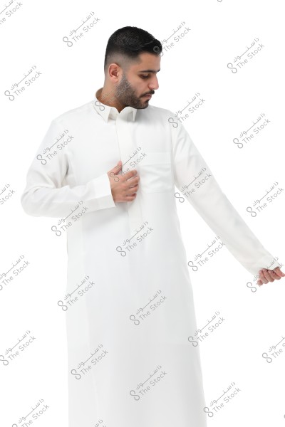 A portrait of a man wearing a traditional white long-sleeve thobe, looking down and to the side, holding one of his sleeves. The thobe resembles traditional clothing from Saudi Arabia and the Gulf region.