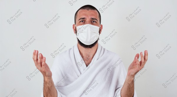 Image of a man wearing a white robe and a white medical face mask, appearing puzzled with his hands slightly raised in a questioning gesture. The background is a neutral white.