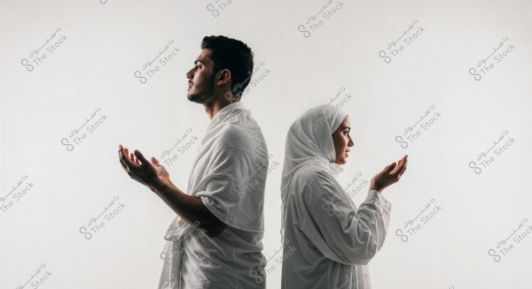 Image of a man and a woman standing back-to-back in traditional white clothing. The man is on the left, with his hands open in prayer, while the woman on the right is wearing a headscarf, also with her hands open in a prayer position. They are dressed in attire associated with Hajj or Umrah rituals. The background is plain white, giving a serene and spiritual atmosphere to the overall scene.
