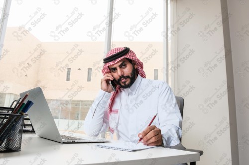 رجل سعودي يرتدي الزي السعودي التقليدي يعمل في مكتب بواجهة زجاجية ويستخدم قلم رصاص لتدوين الملاحظات