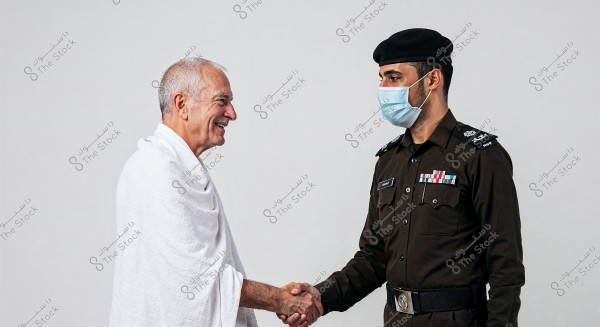 Image of two men shaking hands. The first man is wearing a white Ihram garment, and the second man appears to be a police officer in a dark brown Saudi uniform with a black hat and a blue face mask. The scene reflects friendliness and positive communication between the individuals against a simple white background.