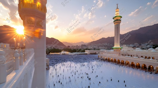 A view of the Prophet\'s Mosque in Medina during sunset, featuring minarets adorned with golden and green lights, and a large gathering of people in the spacious courtyard. The image is centered with mountains and buildings in the background, with clouds lit up in sunset colors in the sky.