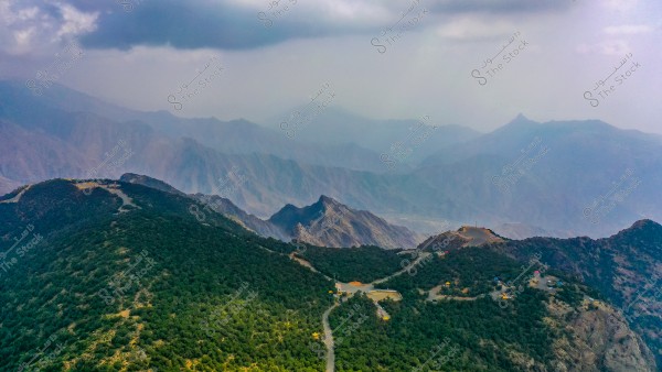 A scenic view of mountainous terrain covered with dense green vegetation under a cloudy sky. In the background, a range of high mountains extends into the horizon, forming overlapping layers of rugged terrain. A few winding roads can also be seen traversing the lush peaks.