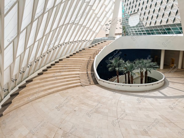 The image depicts a modern interior design of a building, featuring a wide curved staircase made of polished marble or stone, with slanted glass walls allowing natural light to enter. Below, there is a cluster of palm trees planted within a circular space. The design combines contemporary architecture with elements of nature.