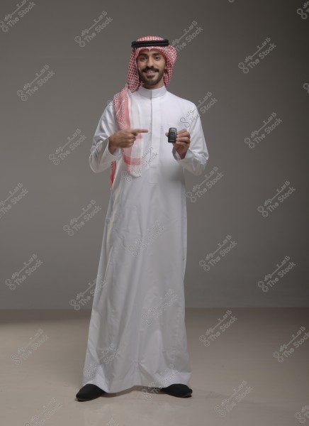 A portrait of a man wearing a traditional white thobe and red checkered keffiyeh, standing in a pose and smiling at the camera. He appears to be from the Gulf region, possibly Saudi Arabia. The man is pointing at something in his hand, which looks like a small electronic device.