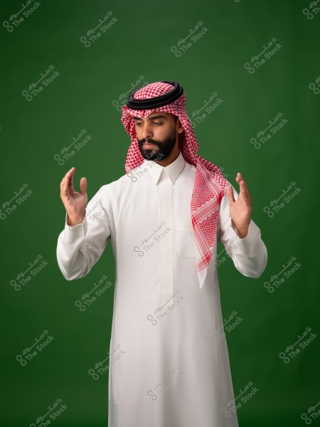 Image of a man wearing traditional Saudi attire, standing against a green background. He is dressed in a white thobe and a red-checkered ghutra and shemagh. He appears to be gesturing with his hands.