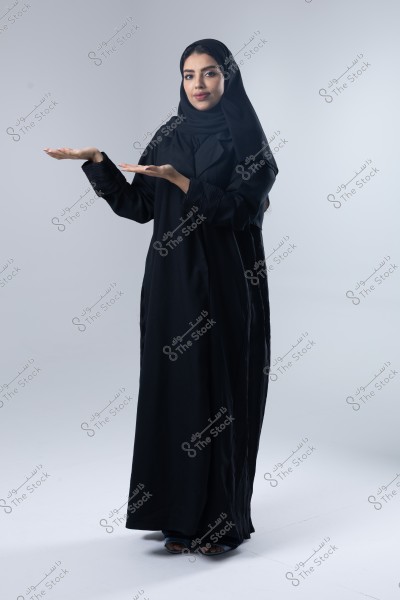 A portrait of a woman wearing a black abaya and hijab, holding her hands in a presenting gesture. The background is white, and she stands upright with a calm expression.