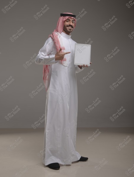 A man wearing traditional Saudi attire, including a thobe, ghutra, and agal, stands smiling in an empty gray studio, holding a tablet in his left hand while pointing to it with his right hand.