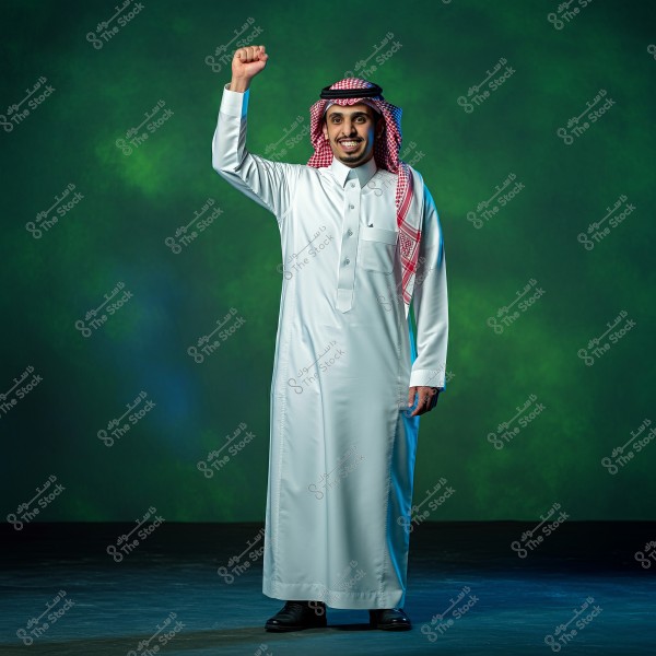 Image of a man wearing traditional Saudi attire, including a white thobe, red ghutra, and agal. The man is standing with a smile and his right hand raised in a gesture of strength or celebration. The background is dark green.