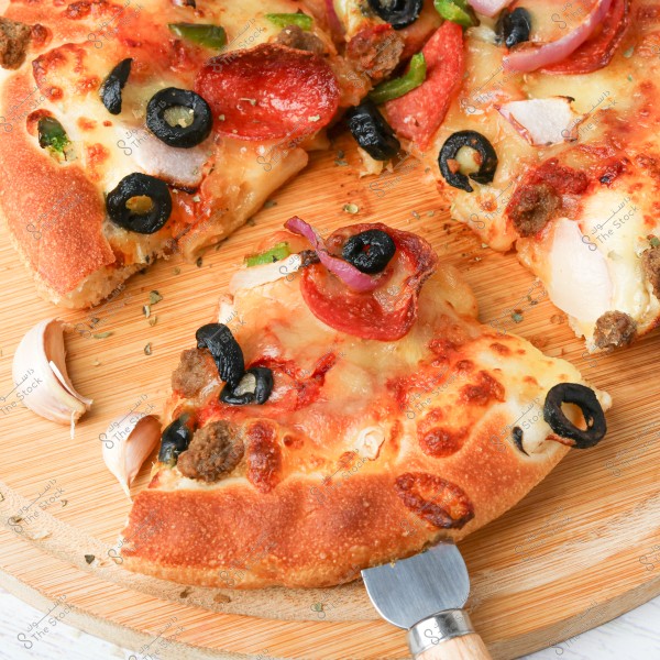 A pizza with slices of pepperoni, black olives, red onion, and melted cheese on a circular wooden board. There are garlic cloves and a sliced piece of pizza with a knife beneath it.
