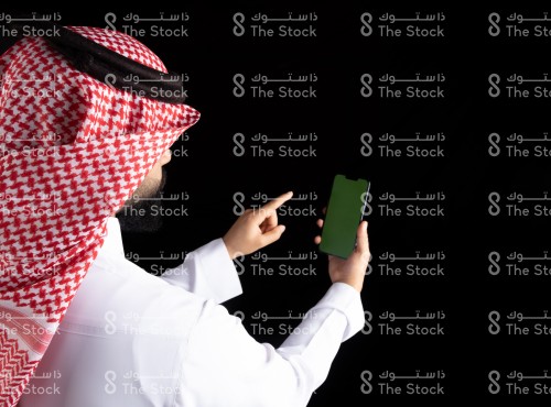 رجل سعودي يرتدي الثوب يحمل الجوال بيده ويشير باصبعه مع خلفية سوداء، رجل يشير الى شيء على الجوال