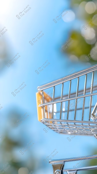 A metallic silver shopping cart with an orange plastic handle, outdoors with a blurred background featuring a blue sky and green bokeh lights.