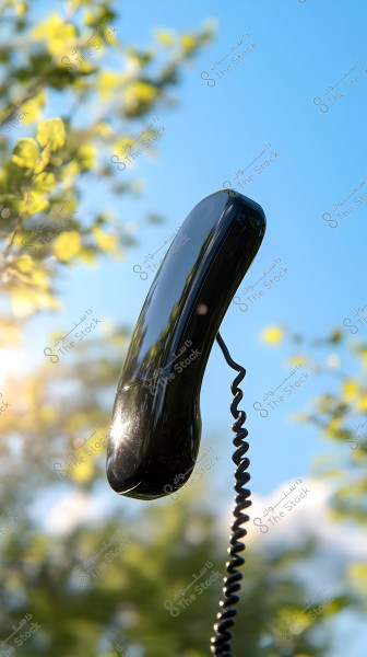An image of a black telephone handset floating in the air against a clear blue sky background. There are green leaves illuminated by sunlight visible on the left side of the image.