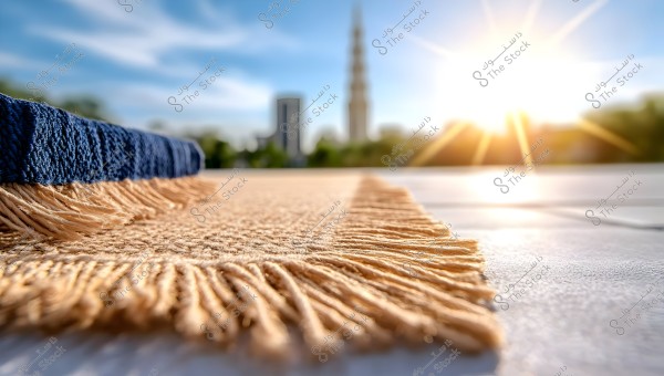 Image shows a prayer rug laid on the ground, focusing on the ornate fringe details. In the background, blurred buildings and green nature appear, with the sun shining in a clear blue sky.