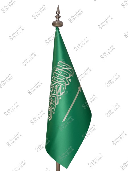 The image shows the flag of Saudi Arabia on a silver pole. The flag is green with the Islamic declaration of faith in Arabic script and a white horizontal sword beneath it.