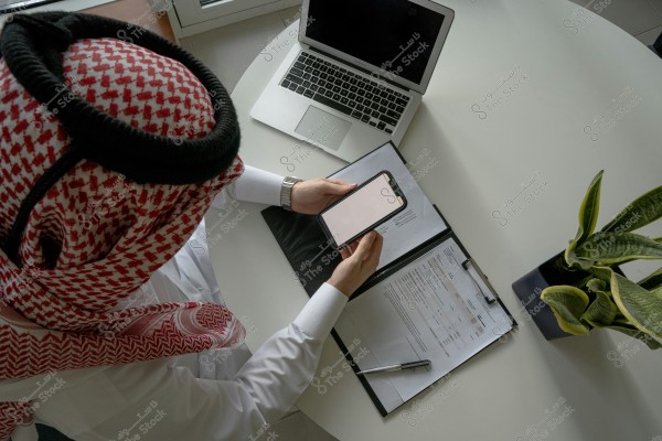 A man wearing traditional Saudi attire, including a white thobe and red shemagh, is seated at a round white table. In front of him is an open laptop and a clipboard with papers and documents, as well as a pen. He is holding a smartphone in his hands. There is a small plant in a pot on the table.