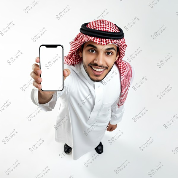 Image of a smiling man wearing traditional attire, including a thobe, ghutra, and agal, likely from the Arabian Peninsula. The man is holding a smartphone with a blank white screen, looking at the camera from a top-down angle. He is dressed in a clean white thobe and black shoes, with a completely white background.