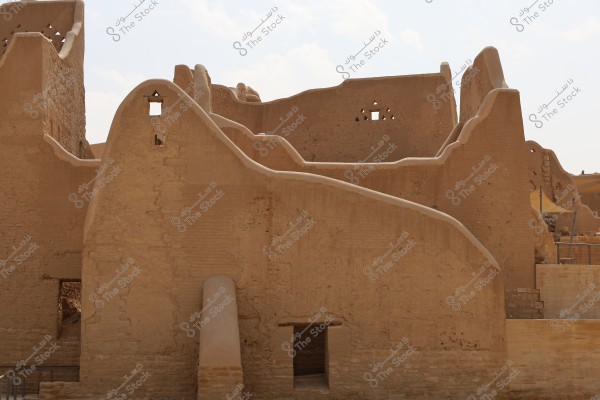 An image showing an old mud-brick building with brown walls and foundations, featuring small square and triangular openings. The building has a wavy design with no roofs. The background sky is partially clear.