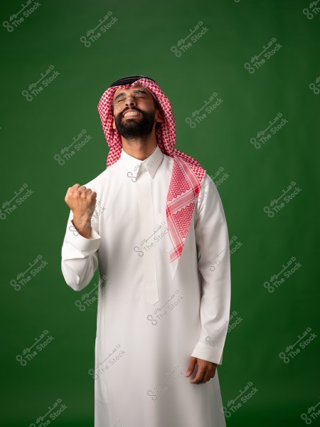 A man wearing a white thobe and a traditional red shemagh stands against a green background. He appears joyful and triumphant.