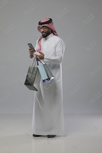 A man stands against a gray background wearing traditional Arabic attire, including a white thobe, a red and white checked headscarf, and a black agal. He is holding several shopping bags and using a smartphone, appearing happy.