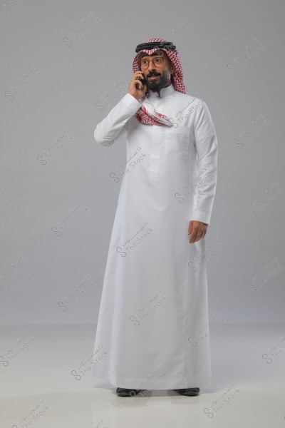 A portrait of a man standing while speaking on the phone. He is wearing a white thobe and a red-checkered headscarf (ghutra) with a black agal, indicating traditional Saudi attire. The man appears to be smiling or engaged in conversation, standing against a simple gray backdrop.