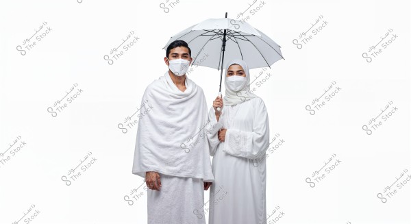 Image of a man and woman wearing Ihram and traditional Islamic attire, both with white medical masks. The woman is holding a white umbrella. The background is completely white.