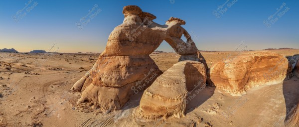 A panoramic image of natural rock formations in the desert under a clear sky. The orange and brown rocks have unconventional shapes, featuring a central rock arch. Light-colored sand surrounds the rocks, with distant hills visible on the horizon.