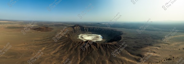 Aerial view of the Al Wahbah volcano crater in the Arabian Peninsula. The crater is prominently circular with white clouds inside it, surrounded by volcanic and mountainous terrain. The background extends toward the horizon with a clear blue sky.