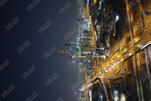 A nighttime view of Riyadh city in Saudi Arabia showing a dense skyline and modern architecture with city lights. The tall buildings are beautifully lit with lights, some featuring unique architectural designs. The highway at the bottom is bustling with traffic, showcasing the bright headlights of the cars.