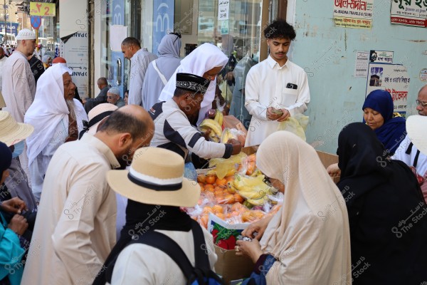 A group of individuals wearing various traditional clothing gather around a fruit stall on the street. A young man in a long Arabic thobe is counting money, while men and women, some wearing hats and abayas, are shopping. The fruits on display include bananas and oranges. Signs in English and Arabic can be seen in the background.