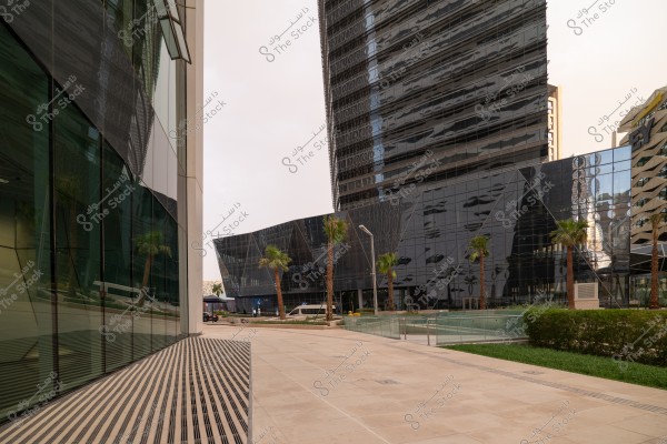 An image depicting a modern urban area with sleek glass architecture, including high and mid-rise glass buildings. The area is adorned with palm trees and surrounding greenery, featuring paved walkways and pedestrian paths. The architecture is contemporary and sophisticated, with excellent lighting reflection.