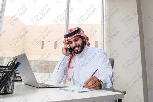 رجل سعودي يرتدي الزي السعودي التقليدي يعمل في مكتب بواجهة زجاجية ويستخدم قلم رصاص لتدوين الملاحظات