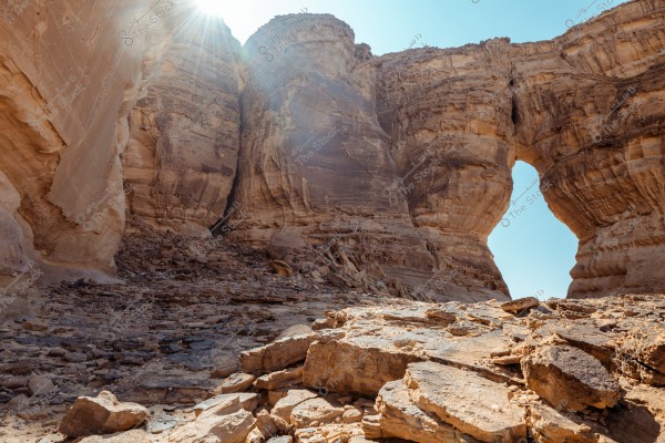 Image of a natural landscape with massive rocks forming a canyon in the desert under a clear blue sky. Sunrays peek through the rocks, casting shadows on the rocky ground below. An arched passage is visible on the right side between the rocks.