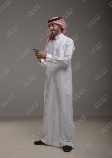 الصورة تظهر رجلًا يقف مرتديًا ثوبًا أبيض مع شماغ أحمر وعقال, وهو يستخدم هاتفًا محمولًا بيديه. الخلفية رمادية بسيطة, ويبدو الرجل مبتسمًا وينظر إلى الهاتف.