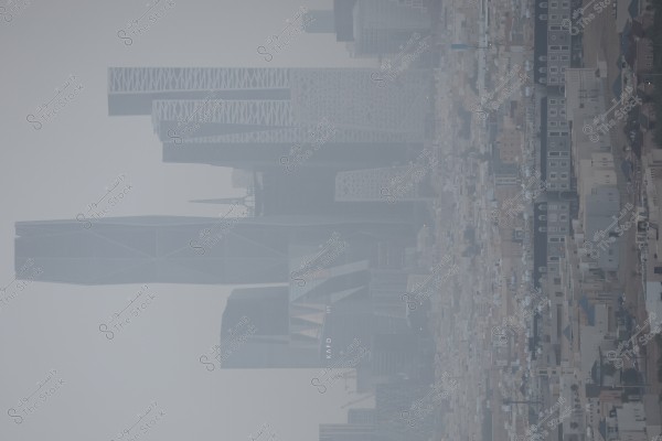 صورة لمشهد حضري في الرياض, المملكة العربية السعودية, تُظهر ناطحات سحاب حديثة مثل مركز الملك عبد الله المالي (KAFD) في يوم ضبابي. تحتل الأبراج ذات التصميمات المعمارية المميزة الجزء العلوي من الصورة, بينما يظهر في الأسفل تفاصيل المباني السكنية التقليدية.