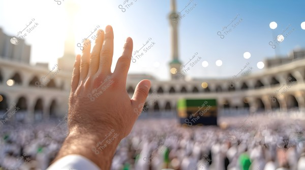 The image shows a hand raised towards the Kaaba in the Grand Mosque in Mecca. The background is blurred, revealing a large crowd of people wearing white clothing, with the upper floor of the mosque visible in the background, along with columns and round lit lights. The weather appears sunny with bright light filling the scene.