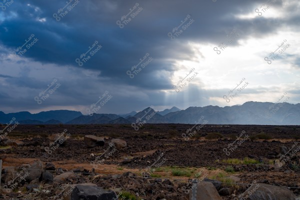A landscape featuring a mountain range under a partially cloudy sky. Sun rays break through the clouds, illuminating the eastern parts of the mountains. The ground in front of the mountains consists of large rocks and dark soil with some scattered green grass.