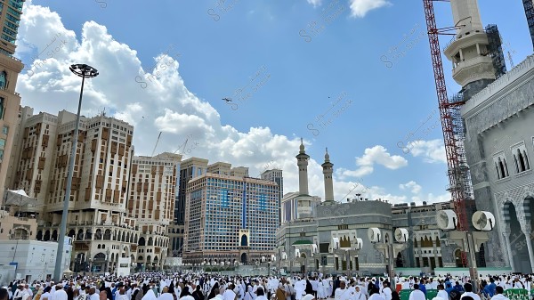 مشهد لمصلين يرتدون الزي الأبيض يتجمعون في ساحة الحرم المكي الشريف مع رؤية مآذن وجزء من الكعبة ومباني متعددة الطوابق في الخلفية, في ظل سماء صافية مع وجود بعض السحب.