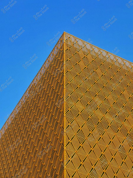 The image depicts a triangular architectural structure with a golden lattice façade composed of an intricate pattern of squares, contrasting against the clear blue sky in the background.