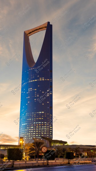 An image of the Kingdom Tower in Riyadh, Saudi Arabia, showcasing its iconic architectural structure with a distinctive curved top. The tower is illuminated with bright lights during sunset, with light clouds in the sky. Some trees and vegetation are visible in the foreground.