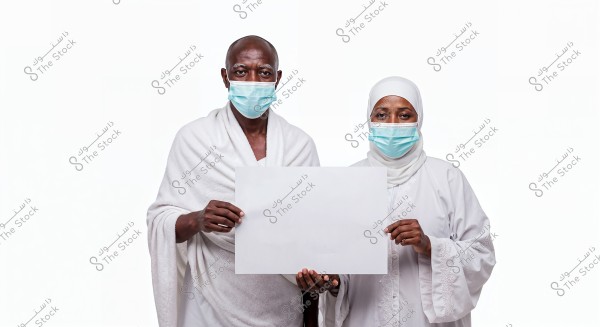 The image shows a man and a woman wearing traditional white Ihram clothing. Both are wearing blue face masks and standing side by side holding a blank white sign. The woman is wearing a headscarf. The image appears to be related to the theme of Hajj or Umrah.