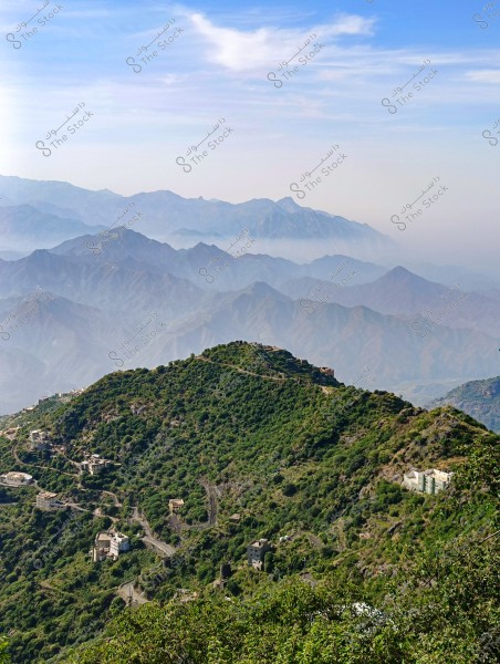 A landscape view of a lush green mountain in the foreground, with small buildings scattered among the trees and winding roads. In the background, a series of cascading mountains are visible, partially covered by mist, under a blue sky with scattered clouds.