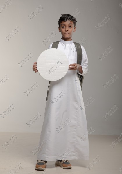 An image of a boy wearing traditional Saudi white attire known as a thobe, carrying a backpack. He is holding a round white board in his hands while standing confidently. He is wearing athletic shoes, and the background is neutral.