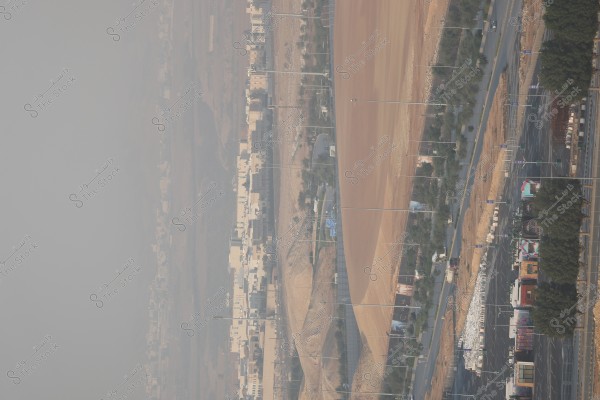 A panoramic view of a city featuring residential buildings with roads and scattered trees. The sky appears slightly hazy, and the street is almost empty of cars. Hills and earthy terrain stand out in the background, presenting a natural and arid appearance.