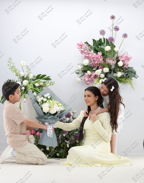 The image shows a woman seated wearing a light yellow dress, with a young girl standing behind her gently embracing her. A boy is kneeling in front of the woman, offering her a bouquet of white and pink flowers. In the background, there is a beautiful arrangement of flowers in various colors such as white, pink, and purple.