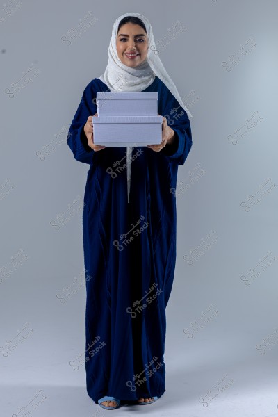 Image of a woman wearing a dark blue abaya and a white hijab, standing and smiling while holding a stack of white boxes. She appears to be from the Gulf countries, with the abaya representing traditional clothing from this region.