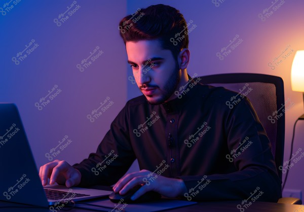 An image of a man sitting in front of a laptop in a dimly lit room with blue and white lighting. He is wearing traditional clothing and appears focused on his screen. A lit lamp on the desk adds to the cozy working atmosphere.